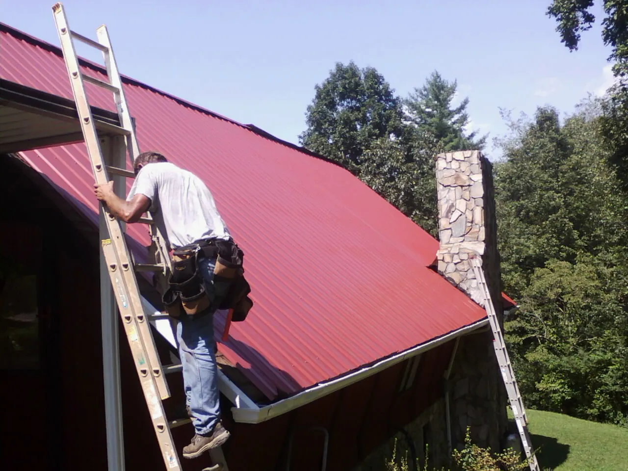 Roofer on a ladder beside a red metal roof and stone chimney — Soffit & Fascia Repair by Roofing Brothers in Etters
